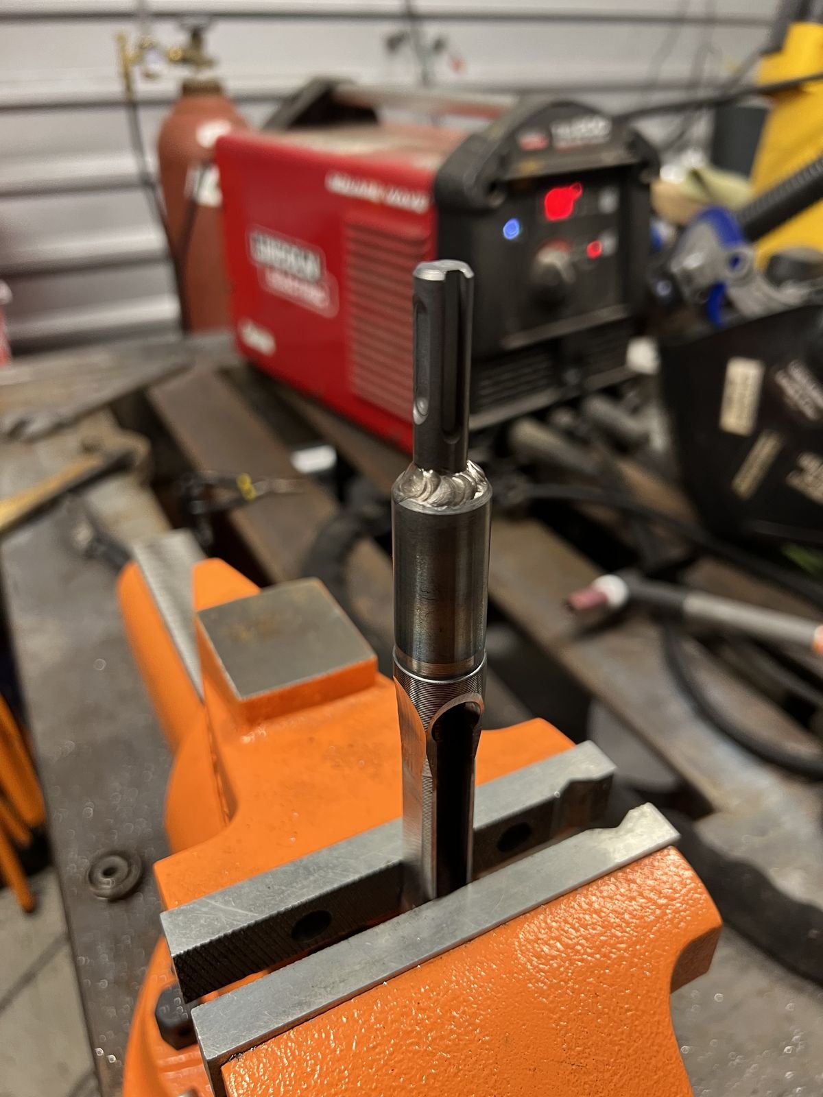 Pony bench vise holding a steel shank part upright, with a Lincoln Electric welder in the background
